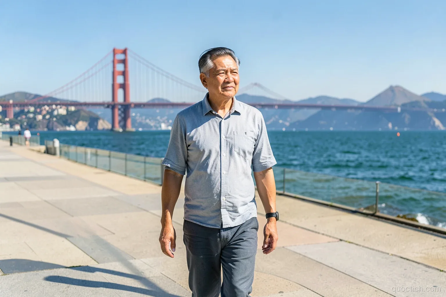 Thoughtful Vietnamese man in his fifties walking alone along a sunny waterfront promenade in San Francisco with the Golden Gate Bridge in the distance, wide shot, clear bright afternoon