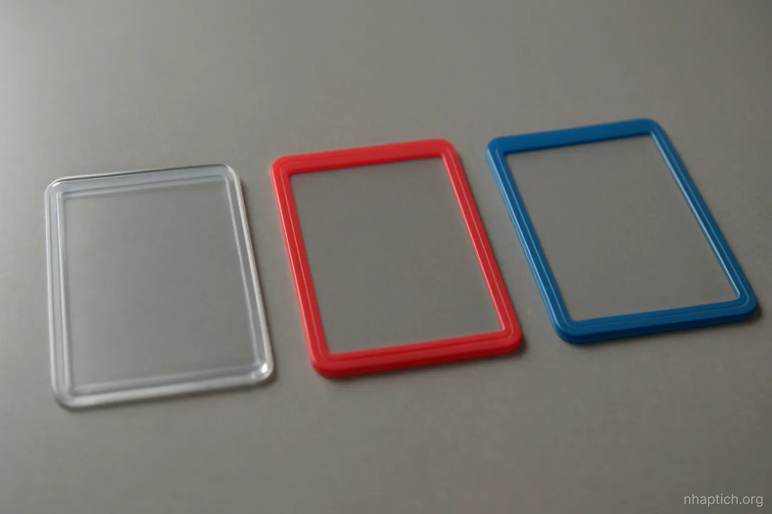 three plain plastic card sleeves of different colors arranged in a row on a grey desk, empty and unmarked